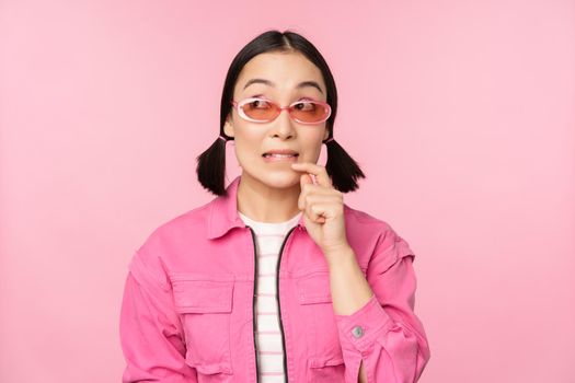 Stylish asian girl in sunglasses, looks thoughtful or intrigued, wants to try smth out, stands over pink background, pondering.