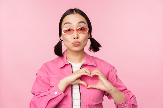 Lovely korean female model in trendy sunglasses, shows heart, care sign, I love you gesture, stands over pink background.