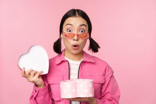 Cute asian girl giving you gift in heart shaped box, kissing and smiling, concept of holiday and celebration, standing over pink background.