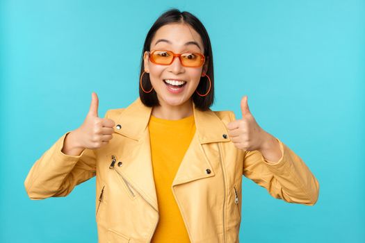 Enthusiastic korean girl looks excited and shows thumbs up, approves smth awesome, excellent choice and quality, stands over blue background.