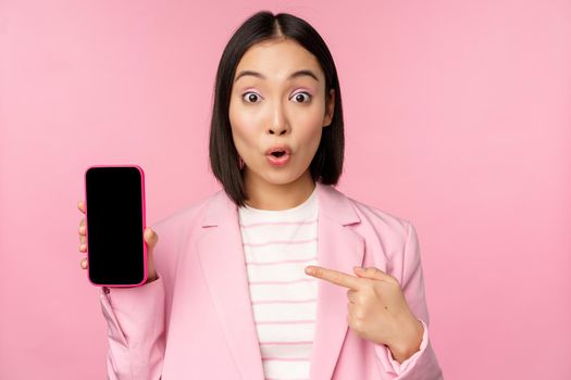 Surprised, enthusiastic asian businesswoman showing mobile phone screen, smartphone app interface, standing against pink background.