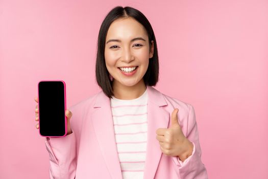 Satisfied smiling asian businesswoman recommending mobile phone app, website company on smartphone, showing screen and thumbs up, pink background.