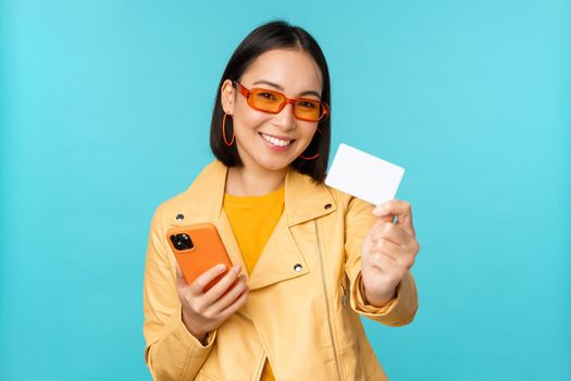 Online shopping. Stylish young asian woman in sunglasses, showing credit card and using smartphone, paying in internet, making purchase, standing over blue background.