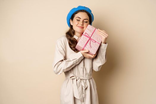 Holidays and gifts concept. Beautiful girl receive gift box and looking happy, holding pink wrapped present with joyful face expression, beige background.