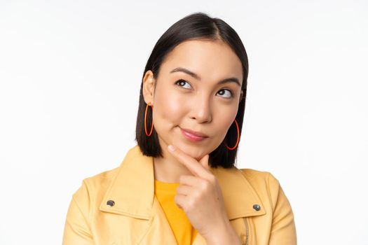 Close up of asian woman thinking, looking up thoughtful, pondering smth with pleased smiling face, standing over white background.