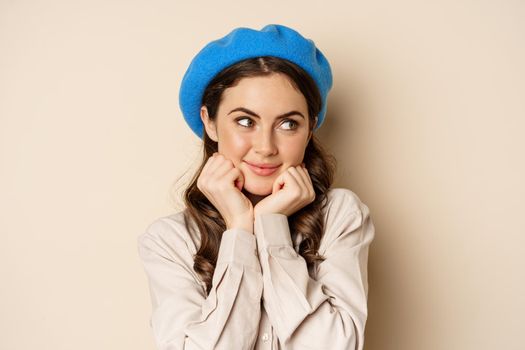 Close up portrait of feminine young woman in trendy french hat, posing cute and romantic, making silly coquettish face and tender smile, beige background.