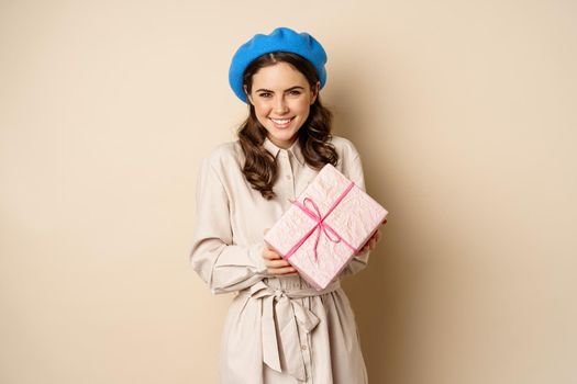 Holidays and gifts concept. Beautiful girl receive gift box and looking happy, holding pink wrapped present with joyful face expression, beige background.