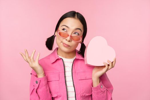 Image of stylish asian girlfriend in sunglasses, guessing whats inside gift box, heart-shaped present, standing over pink background.