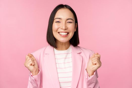 Enthusiastic saleswoman, asian corporate woman say yes, achieve goal and celebrating, triumphing, looking with rejoice and smiling, standing over pink background.