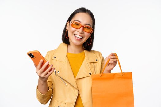 Stylish asian girl shopper, wears sunglasses, holding shopping bag and smartphone, going for discounts in stores, standing over white background.