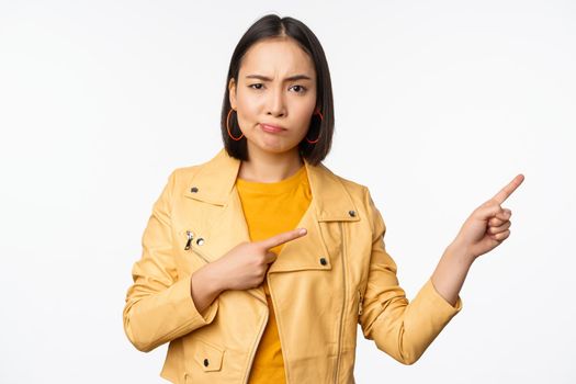 Disappointed asian girl frowning, sulking upset, pointing fingers right at advertisement, standing over white background. Copy space