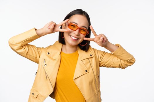 Portrait of stylish asian modern girl, wearing sunglasses and yellow jacket, showing peace, v-sign gesture, standing over white background, happy smiling face.