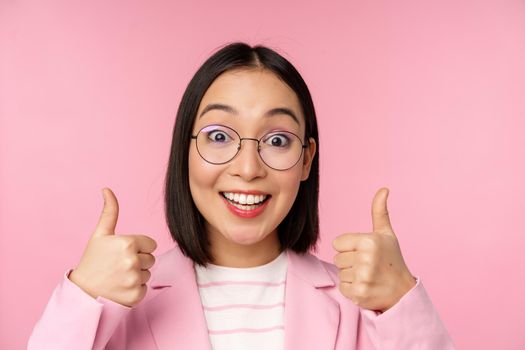 Awesome, congrats. Face of excited asian businesswoman in glasses, smiling pleased, showing thumbs up in approval, standing over pink background.