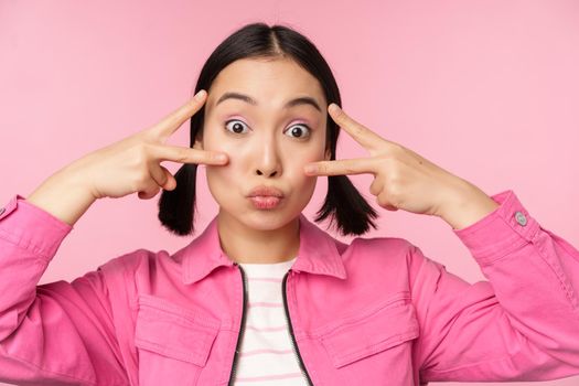 Close up of stylish asian girl smiles happy, shows peace v-sign, kawaii pose, posing against pink background. Copy space