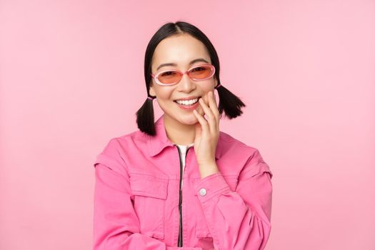 Close up portrait of stylish asian woman in sunglasses, smiling, looking flattered, laughing coquettish, standing over pink background. Copy space