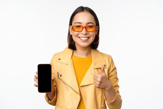 Beautiful korean girl, asian woman in sunglasses, showing smartphone app interface, thumbs up, recommending mobile phone application, white background.