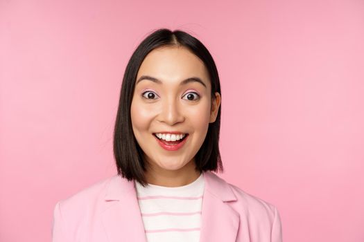 Close up portrait of asian corporate woman, business lady looking surprised and amazed at camera, standing in suit against pink background.