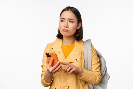 Image of happy girl traveller, tourist with backpack looking at smartphone, using mobile phone route app, standing over white background. Copy space