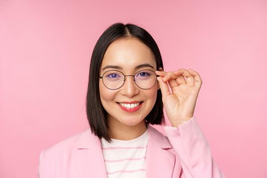 Close up portrait of asian corporate woman, professional businesswoman in glasses, smiling and looking confident at camera, pink background.