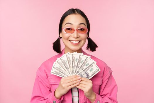 Microcredit and fast loans concept. Excited stylish korean girl, showing money, cash dollars and looking happy, standing in sunglasses over pink background.