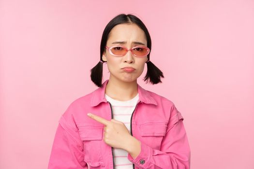 Potrait of cute korean girl in sunglasses, pointing left and looking disappointed, sulking upset, standing over pink background.