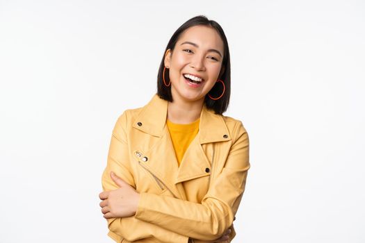 Image of beautiful modern asian girl laughing, smiling and looking happy at camera, standing in yellow jacket against white background. people concept