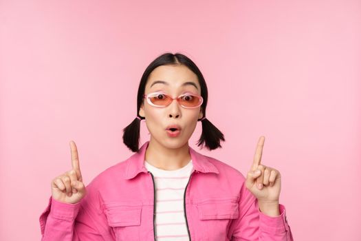 Close up portrait of asian woman gasping, looking surprised, pointing finger at banner, advertisement, standing over pink background.