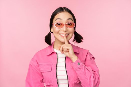 Modern korean girl in stylish spring outfit, sunglasses, showing shush, hush sign, press finger to lips, taboo gesture, standing over pink background.