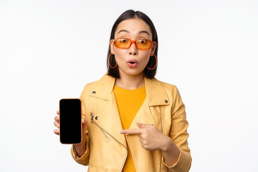 Portrait of stylish korean girl in sunglasses, smiling, pointing finger at smartphone screen, showing mobile phone application, standing over white background.