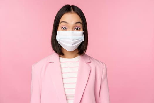 Portrait of asian businesswoman in medical face mask, wearing suit, concept of office work during covid-19 pandemic, standing over pink background.
