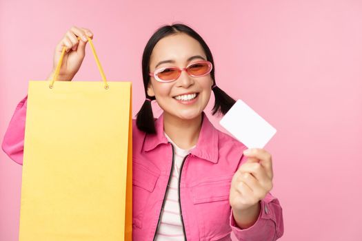 Happy young asian woman showing credit card for shopping, holding bag, buying on sale, going to the shop, store, standing over pink background.