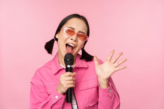 Happy beautiful asian girl singing with mic, using microphone, enjoying karaoke, posing against pink studio background.