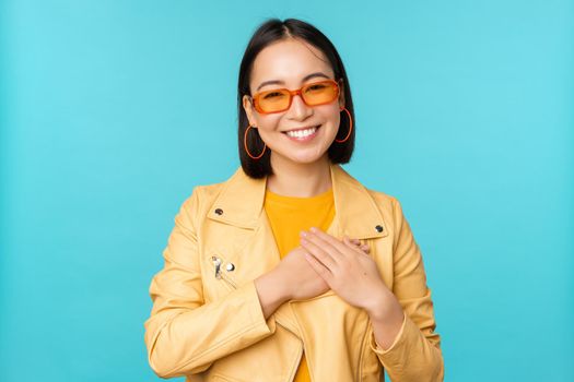 Portrait of asian woman smiling, holding hands on heart and looking with tenderness, care at camera, thankful emotion, standing over blue background.