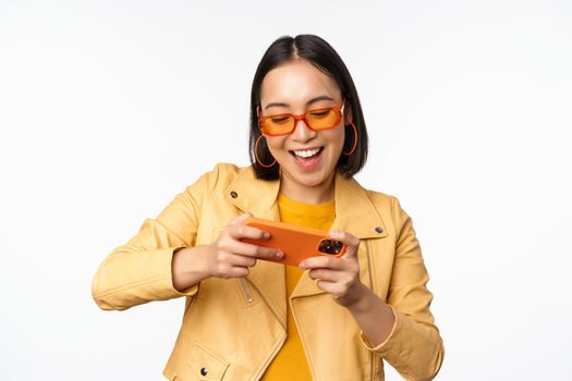 Stylish korean girl in sunglasses, playing mobile video game, laughing and smiling while using smartphone, standing over white background.