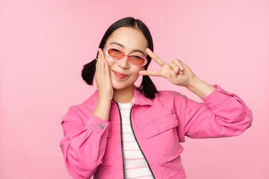 Portrait of kawaii asian girl in sunglasses, showing peace, v-sign near eye and smiles cute at camera, posing in trendy clothes against pink background.