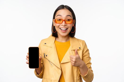 Beautiful korean girl, asian woman in sunglasses, showing smartphone app interface, thumbs up, recommending mobile phone application, white background.