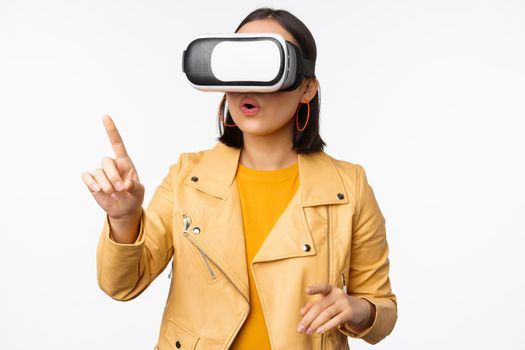 Young asian girl wearing VR glasses, looking amazed, using virtual reality headset, posing against white background.