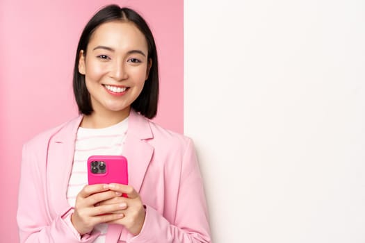 Image of korean female entrepreneur in suit, standing near info wall, advertisement on board, holding smartphone and smiling, posing over pink background.