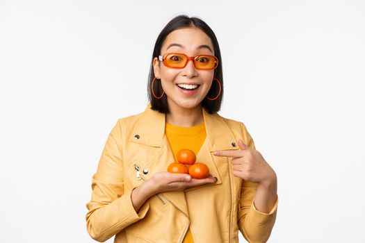 Stylish happy asian girl in sunglasses holding tangerines and smiling, posing against white background. Copy space