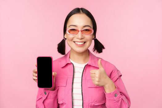 Happy stylish girl recommends application on mobile phone. Smiling asian female model showing smartphone screen and thumb up, standing over pink background.