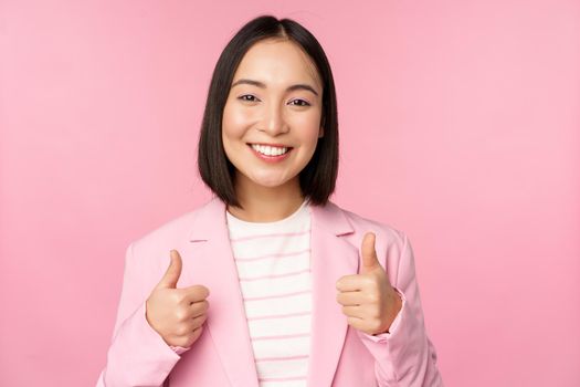 Professional businesswoman, asian corporate woman showing thumbs up and smiling, praise and compliment, standing in suit over pink background.