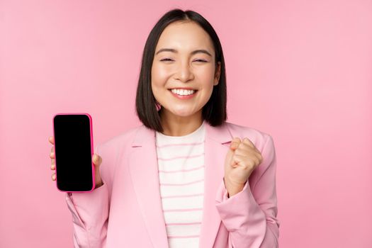 Winning korean businesswoman showing smartphone screen, smiling pleased, demonstrating mobile phone application, online store or shopping app, standing over pink background.