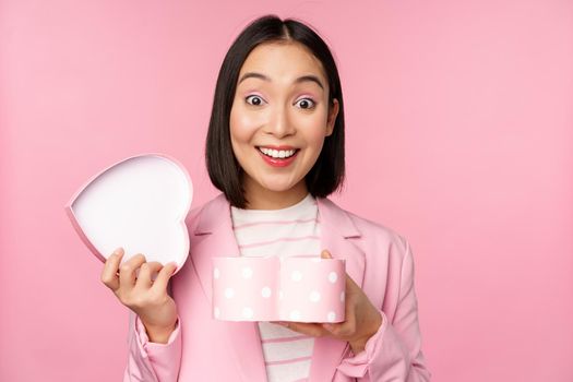 Happy cute korean girl in suit, opens up heart shaped box with romantic gift on white day holiday, standing in suit over pink background.