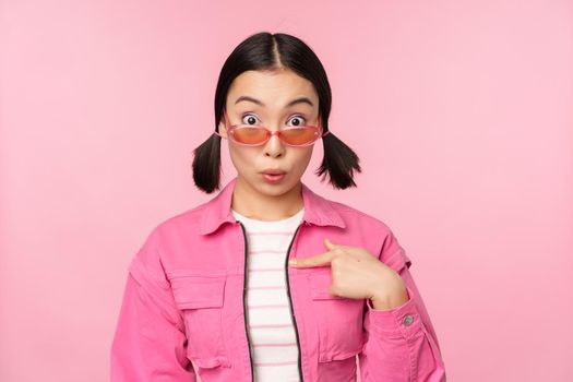 Close up portrait of asian girl looks surprised, points at herself with disbelief, being chosen, stands over pink background. Copy space