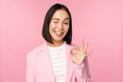 Excellent work, well done gesture. Smiling asian businesswoman showing okay, ok sign, praise good work, recommending company, looking as confident professional, pink background.