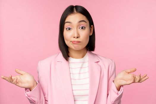 Portrait of confused asian businesswoman shrugging shoulders, looking clueless and puzzled, dont know, cant say, standing over pink background in office suit.