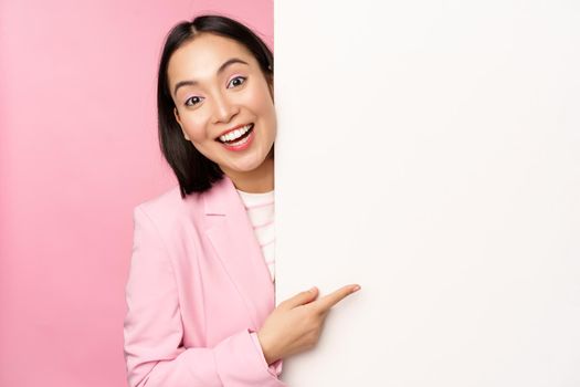 Portrait of young japanese business woman, corporate lady in suit pointing on wall with chart, showing diagram or advertisement on empty copy space, pink background.