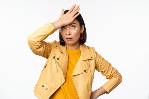 Annoyed and tired asian girl, slap forehead, facepalm and eyeroll with bothered face expression, standing in casual clothes over white studio background.