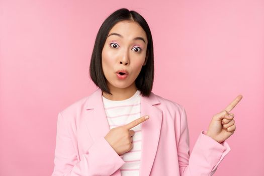 Enthusiastic professional businesswoman, saleswoman pointing fingers right, showing advertisement or company logo aside, posing over pink background.