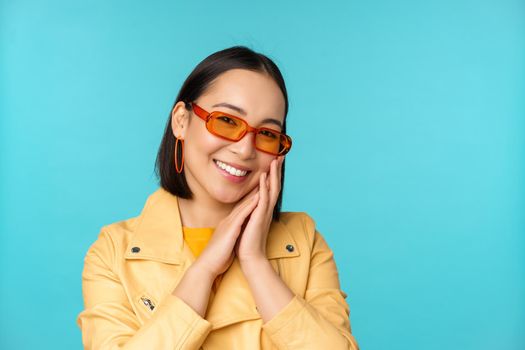 Close up portrait of asian young woman in sunglasses, smiling and looking romantic, standing happy over blue background. Copy space